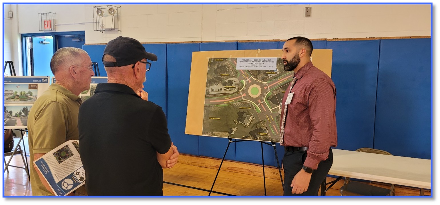 "Three individuals are standing in a gymnasium near a large display board mounted on an easel. The board shows a detailed diagram of a proposed roundabout with labeled roadways and landscaped areas. One individual is standing beside the board and appears to be explaining the diagram to the other two individuals, who are viewing the display. Blue padded wall panels, gym flooring, and an exit door are visible in the background."